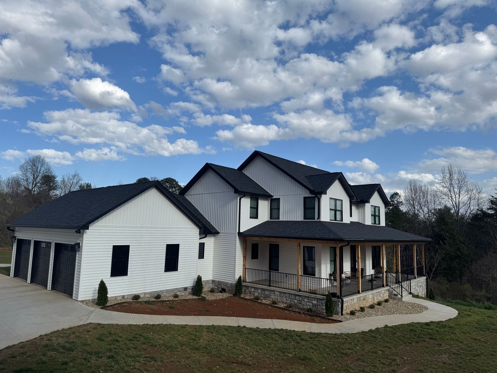 White two-story house with black roof, front porch, and three-car garage under a cloudy blue sky