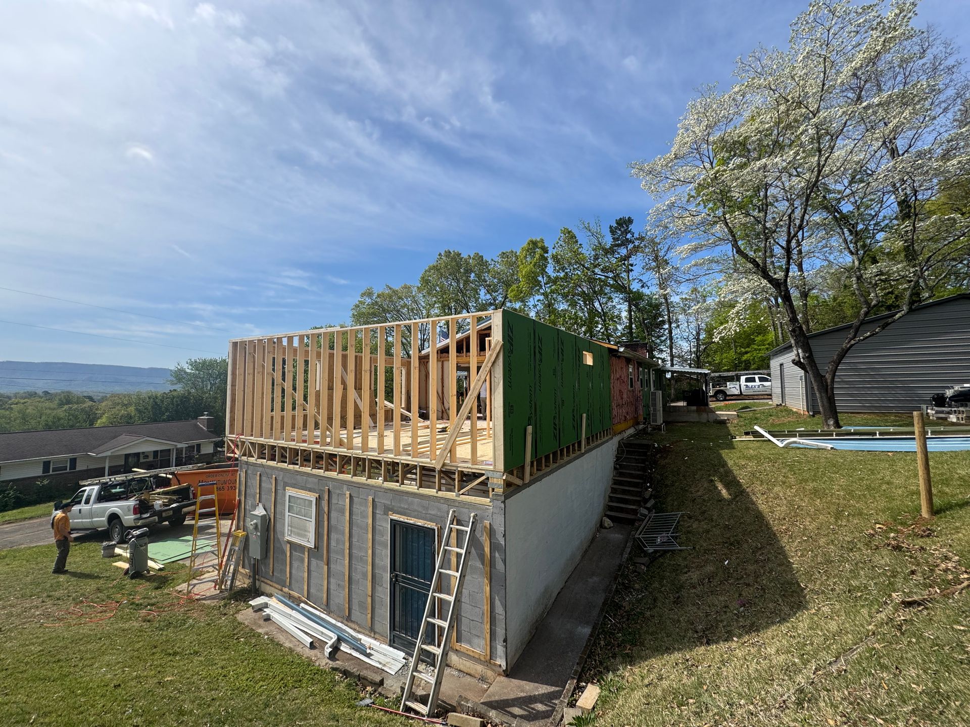Partially built house with exposed wood framing and a gray lower level on a grassy hillside under a blue sky