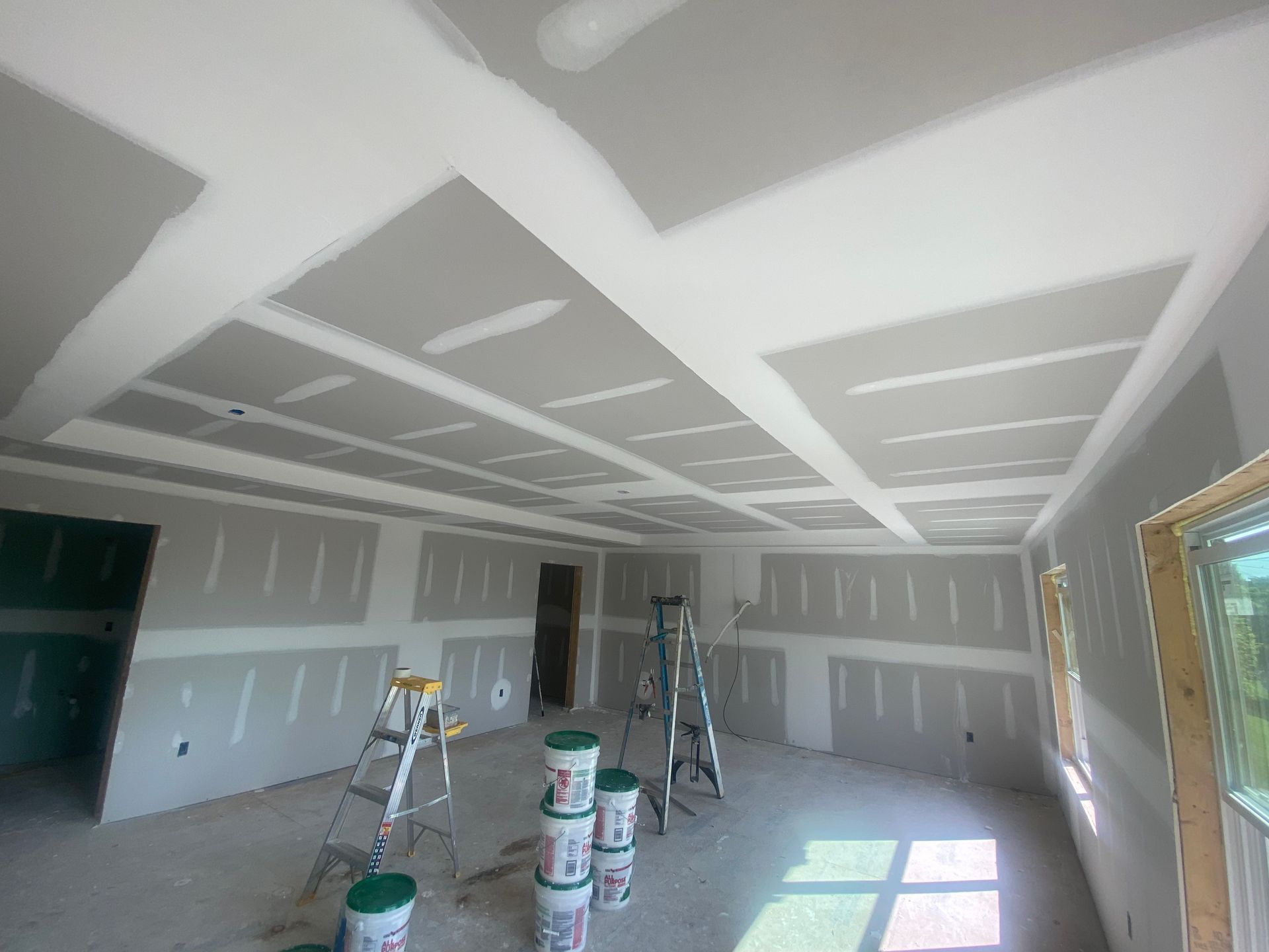Drywalled interior room under construction with ladders, paint buckets, and sunlight on the floor.