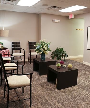 Waiting room with neutral walls, several chairs, and small tables with plants. A door marked