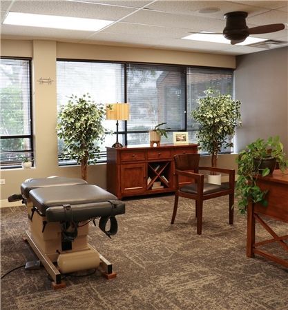 Chiropractor's office with an adjusting table, wooden cabinet, chairs, and plants near a large window with blinds. The room has neutral colors.