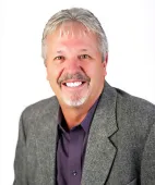 A person with short, light-brown hair and a mustache smiling in a purple button-down shirt and a grey textured blazer.