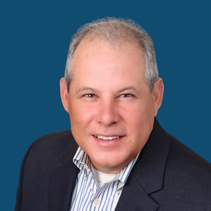 Smiling man in a navy blazer over a striped shirt against a blue background.
