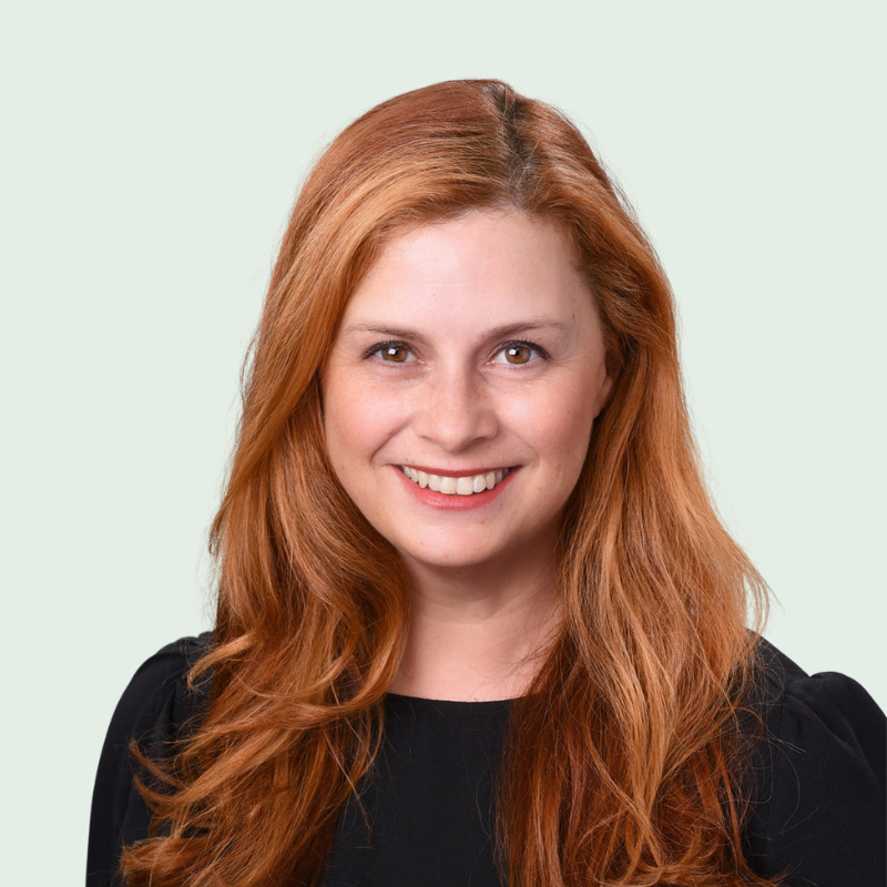 Woman with long red hair smiles in front of a light green background. She is wearing a black top.