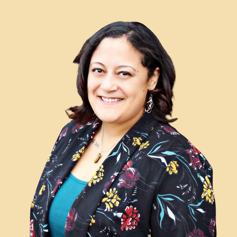 Woman with dark hair smiles, wearing a floral blazer over a teal top, against a yellow background.
