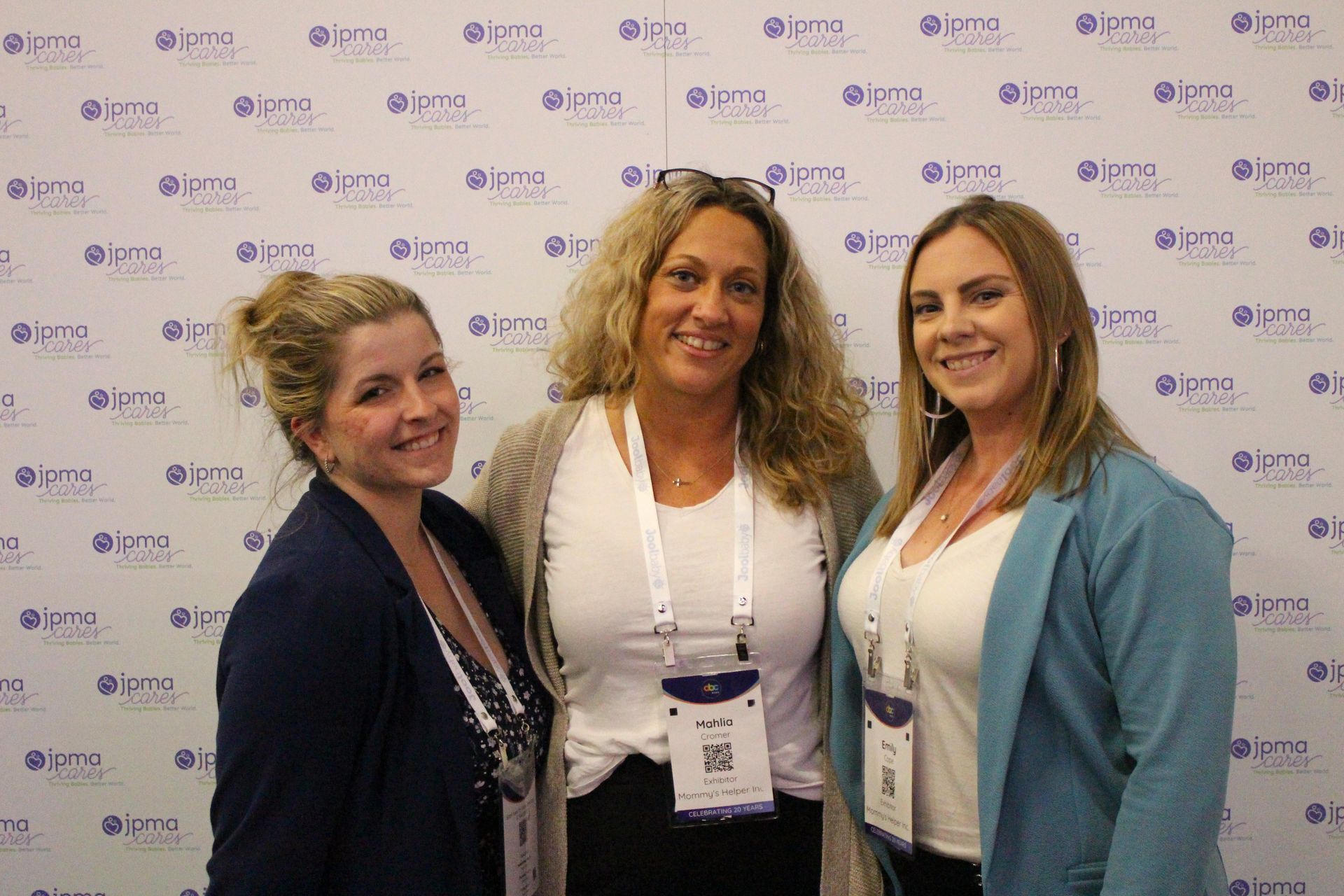 Three women smiling, posing in front of a backdrop with a logo; one in blue, one in gray, and one in a teal blazer.