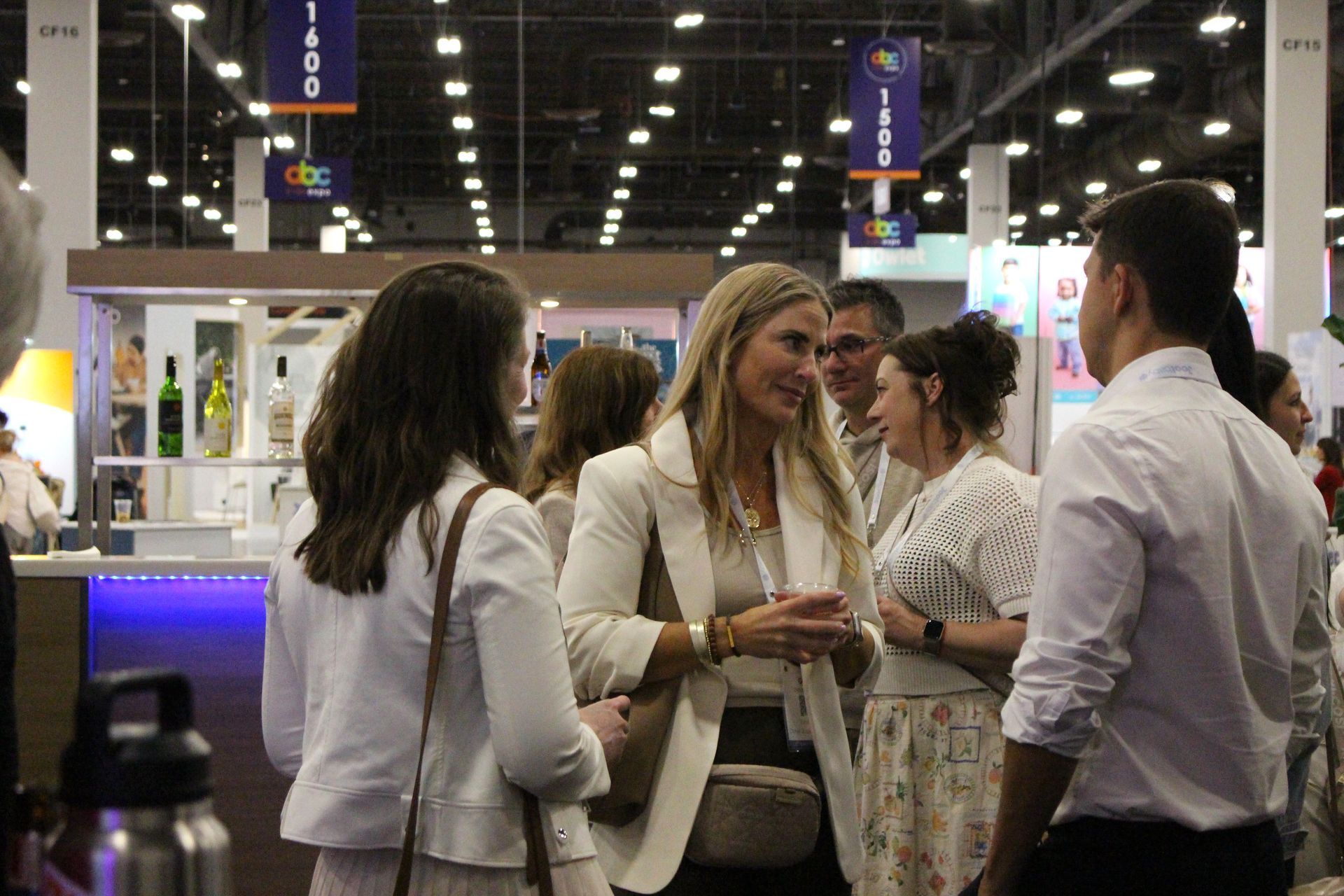 People talking at a trade show. Blonde woman in white blazer gestures while speaking. Bright overhead lights.
