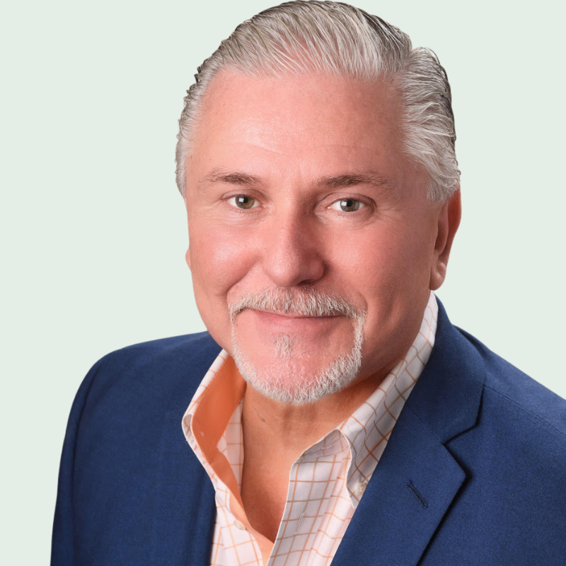 Man with gray hair and beard, wearing a blue blazer and orange shirt, smiling.