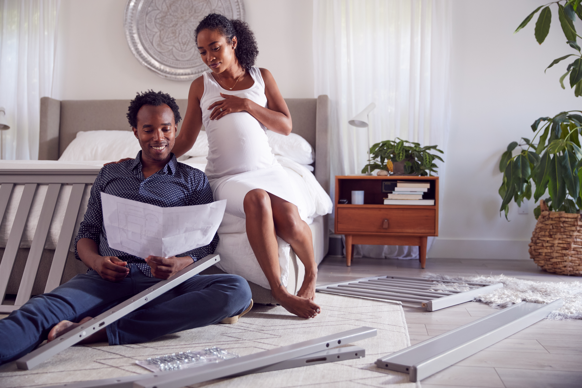Couple assembling a crib in a bedroom. Pregnant woman touches her belly while the man reads instructions.