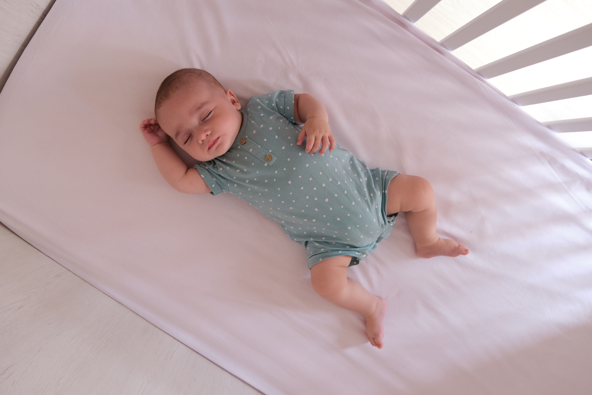 Baby sleeping on a white sheet in a crib, wearing a green jumpsuit representing safe sleep environment.