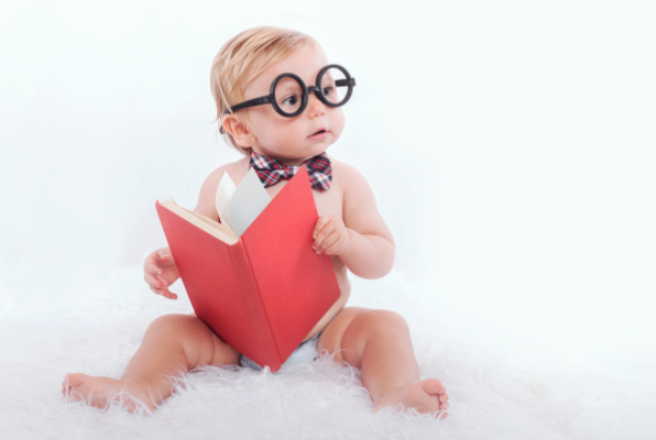 Baby wearing glasses and bow tie, holding a red book, sitting on white fluffy surface.