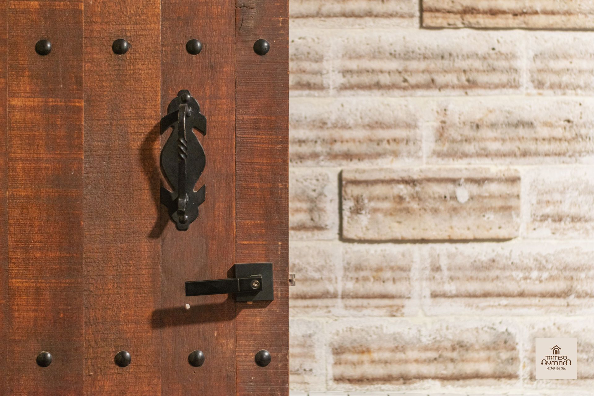 Primer plano de una puerta de madera con tirador de hierro negro junto a un muro de piedra clara.