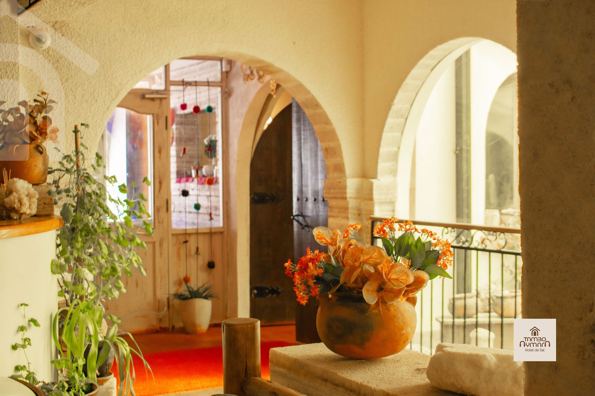 Pasillo soleado con arcos, plantas en macetas y flores naranjas sobre una mesa de piedra.