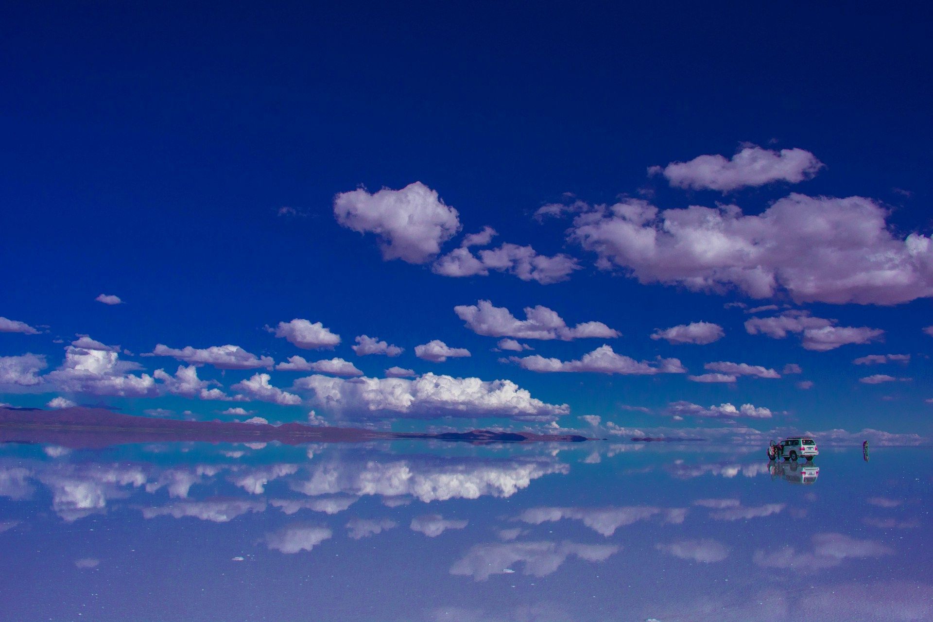 Cielo azul con nubes blancas dispersas reflejadas en aguas tranquilas, con un pequeño árbol a la derecha.