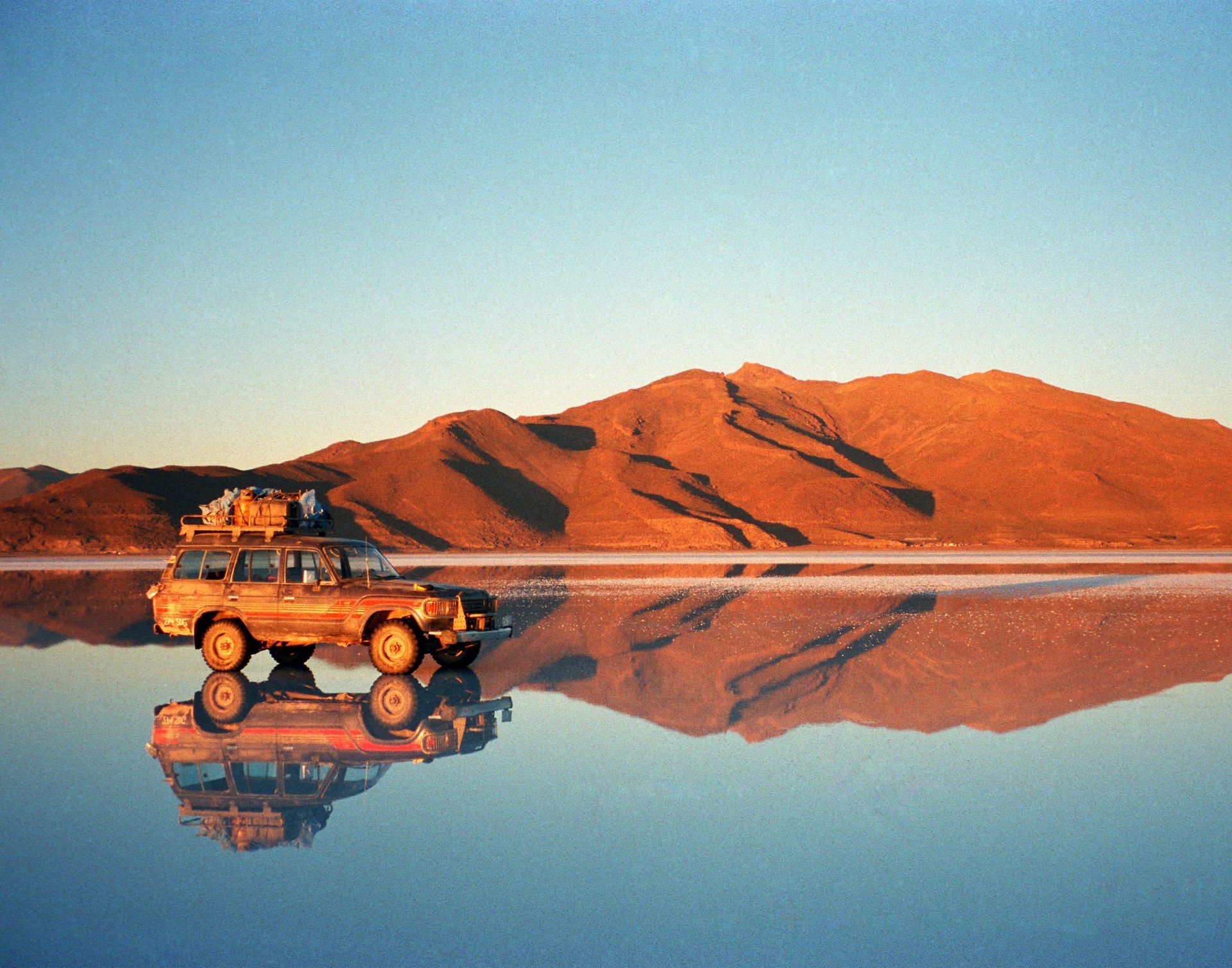 Vehículo todoterreno en una llanura salina reflectante con montañas rojas y cielo azul al amanecer.