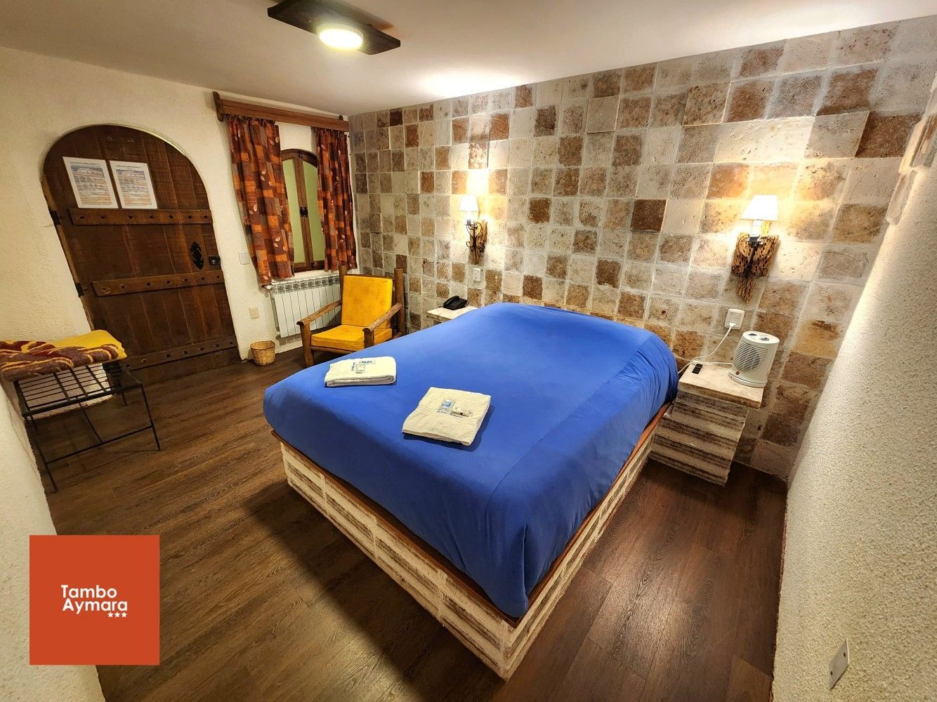 Habitación acogedora con colcha azul, muebles de madera, pared estampada e iluminación cálida.