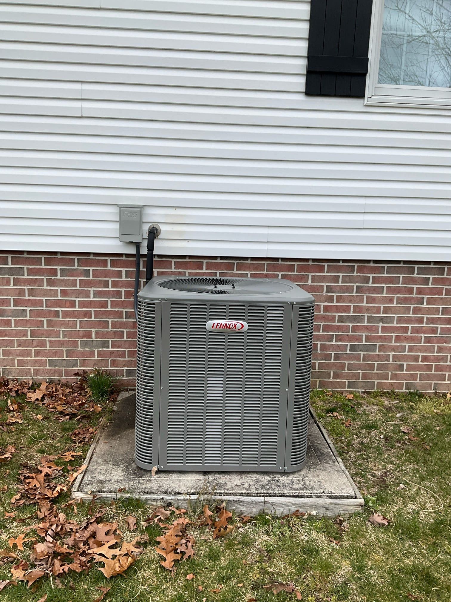 Gray HVAC unit beside a house with white siding and red brick foundation on a concrete pad