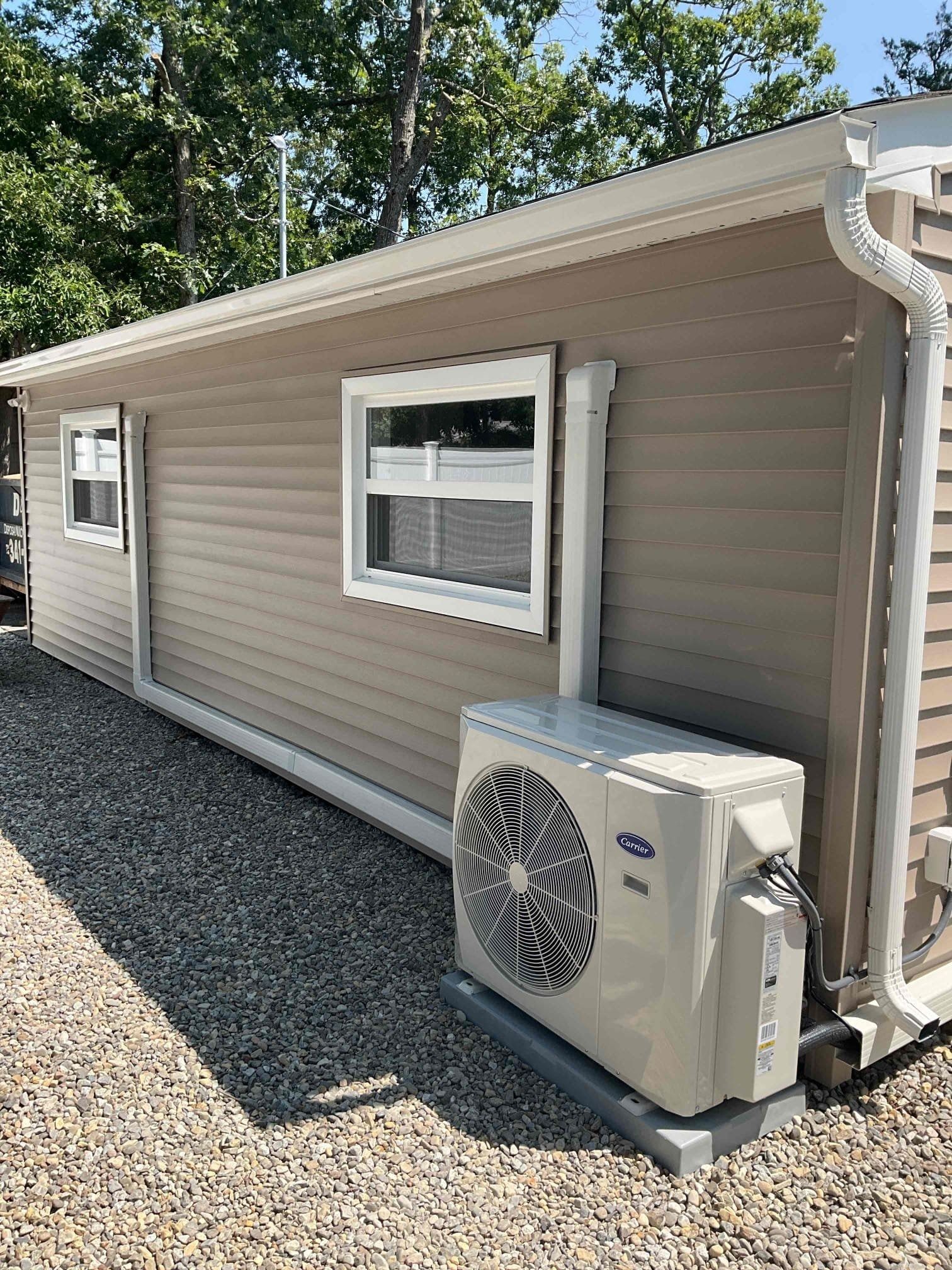 Single-story beige house exterior with white-trim windows and a wall-mounted air conditioner unit on gravel.
