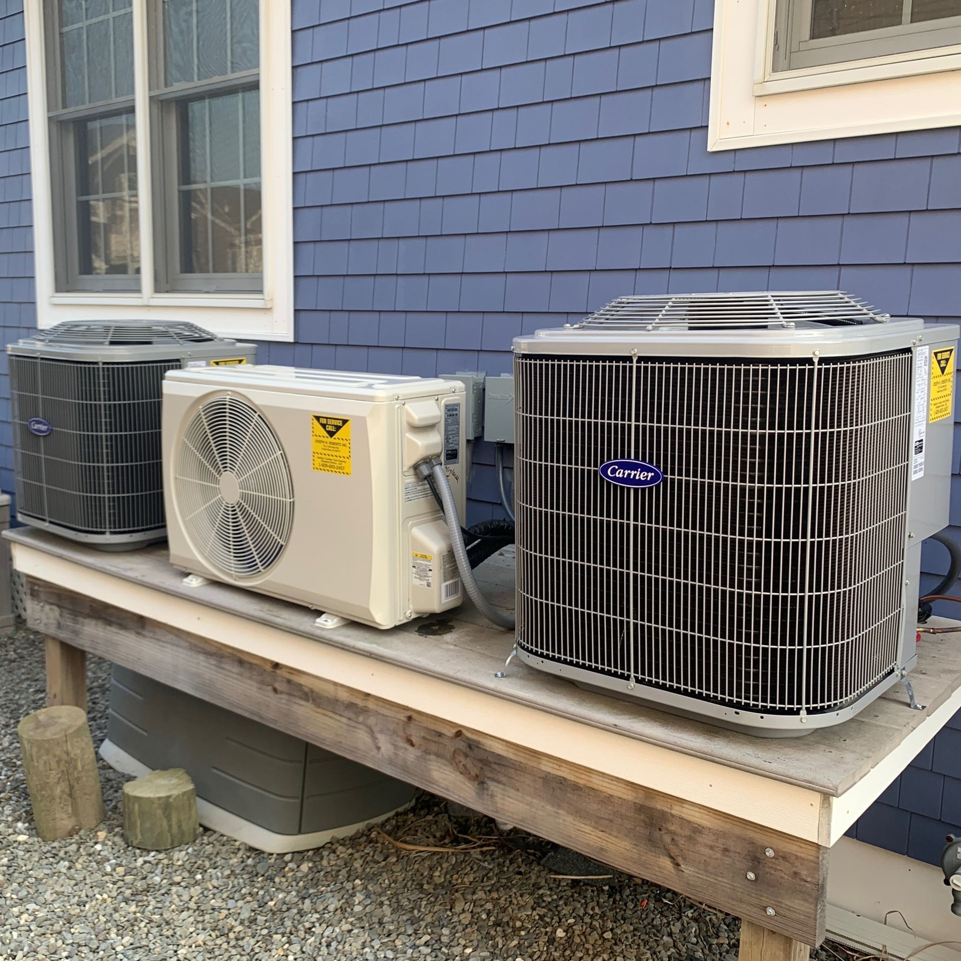 Three outdoor HVAC units on a wooden platform beside a blue house.