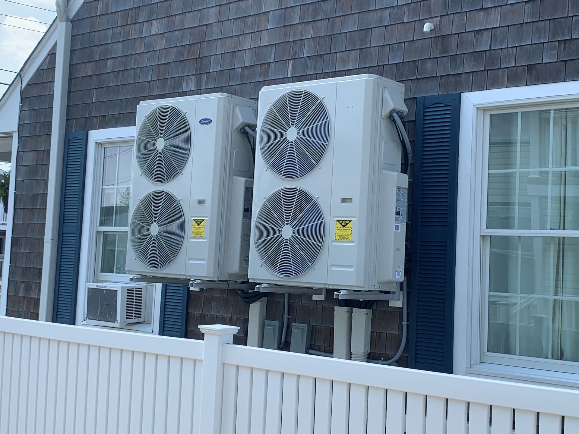 Two outdoor air conditioning units mounted on a house exterior beside a window and white fence