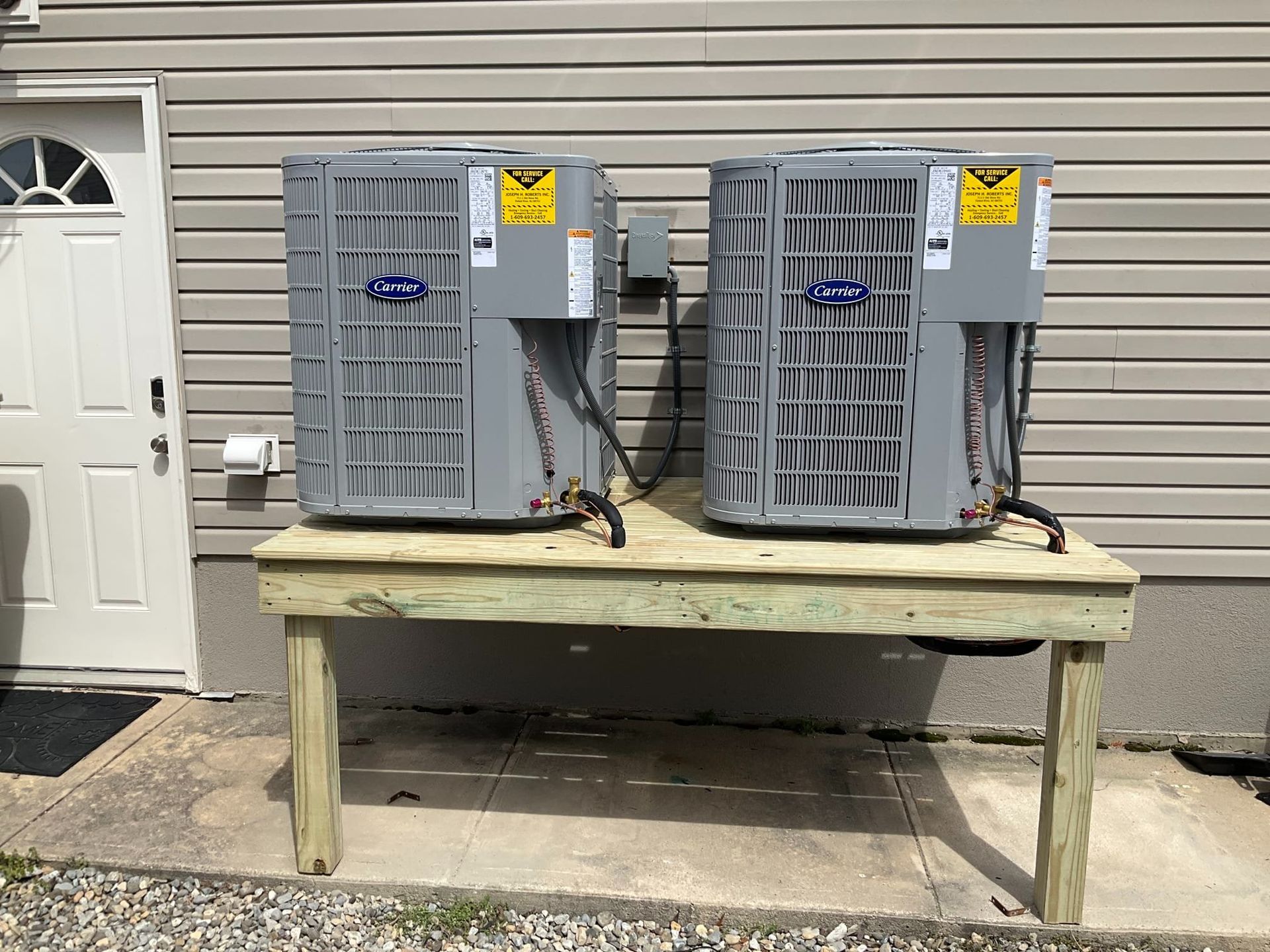 Two gray HVAC units on a wooden platform beside a house door and wall.