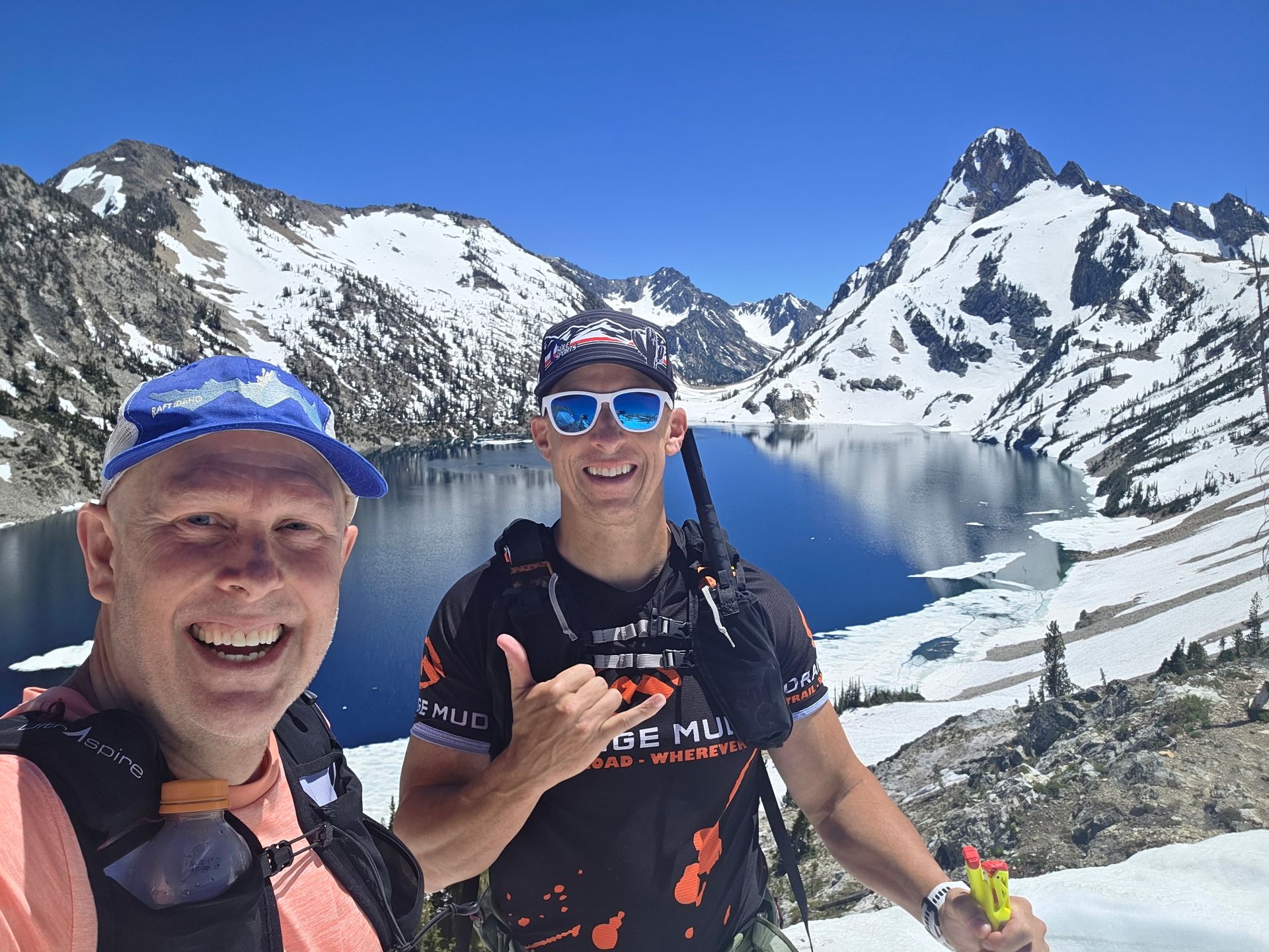 Two people hike on a mountain ridge with lush green vegetation. One takes a selfie; blue sky.