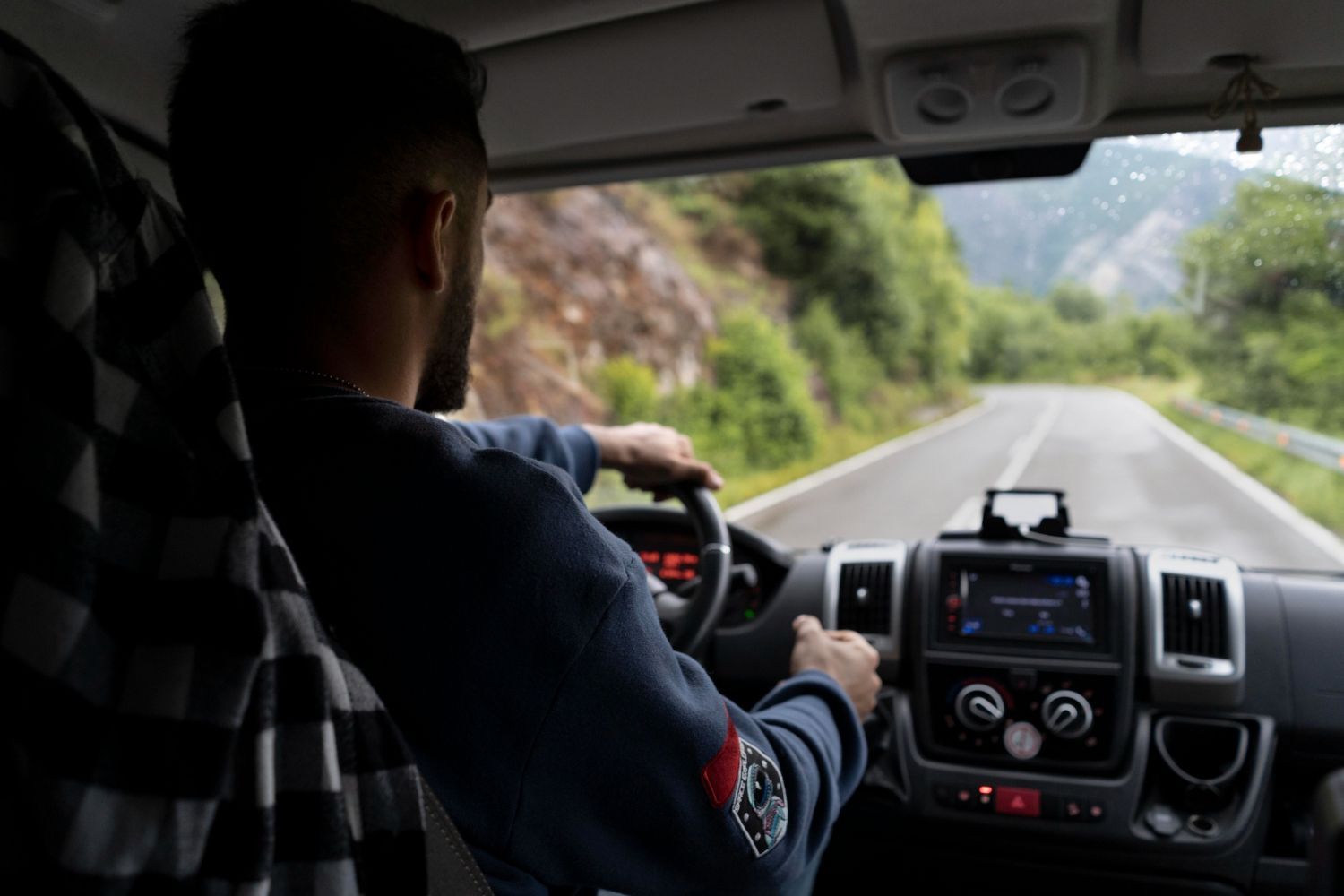 Driver steering on a mountain road, seen from inside a car with a checkered passenger seat cover.