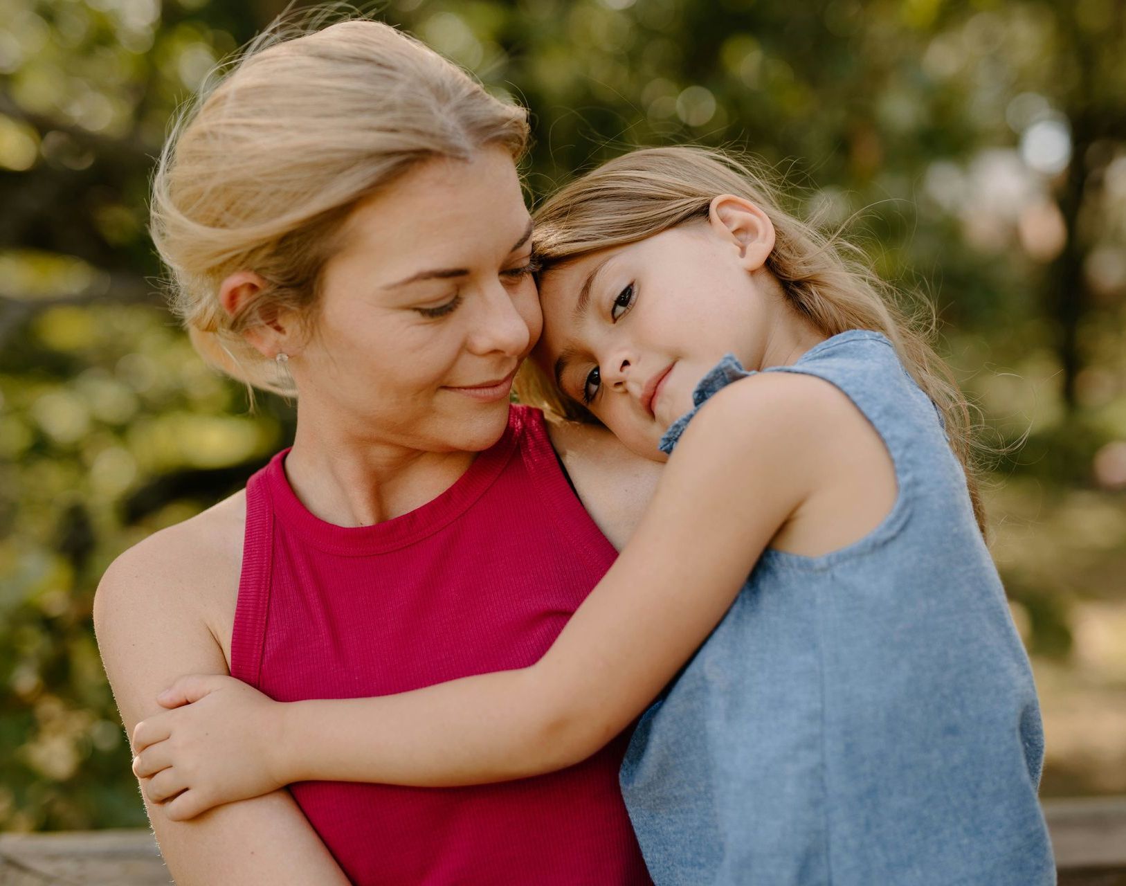 Woman in a pink top hugging a child in a blue shirt outdoors, both resting their heads together.