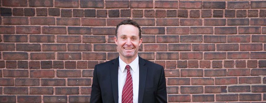 Man in a suit and red tie standing against a brick wall, smiling