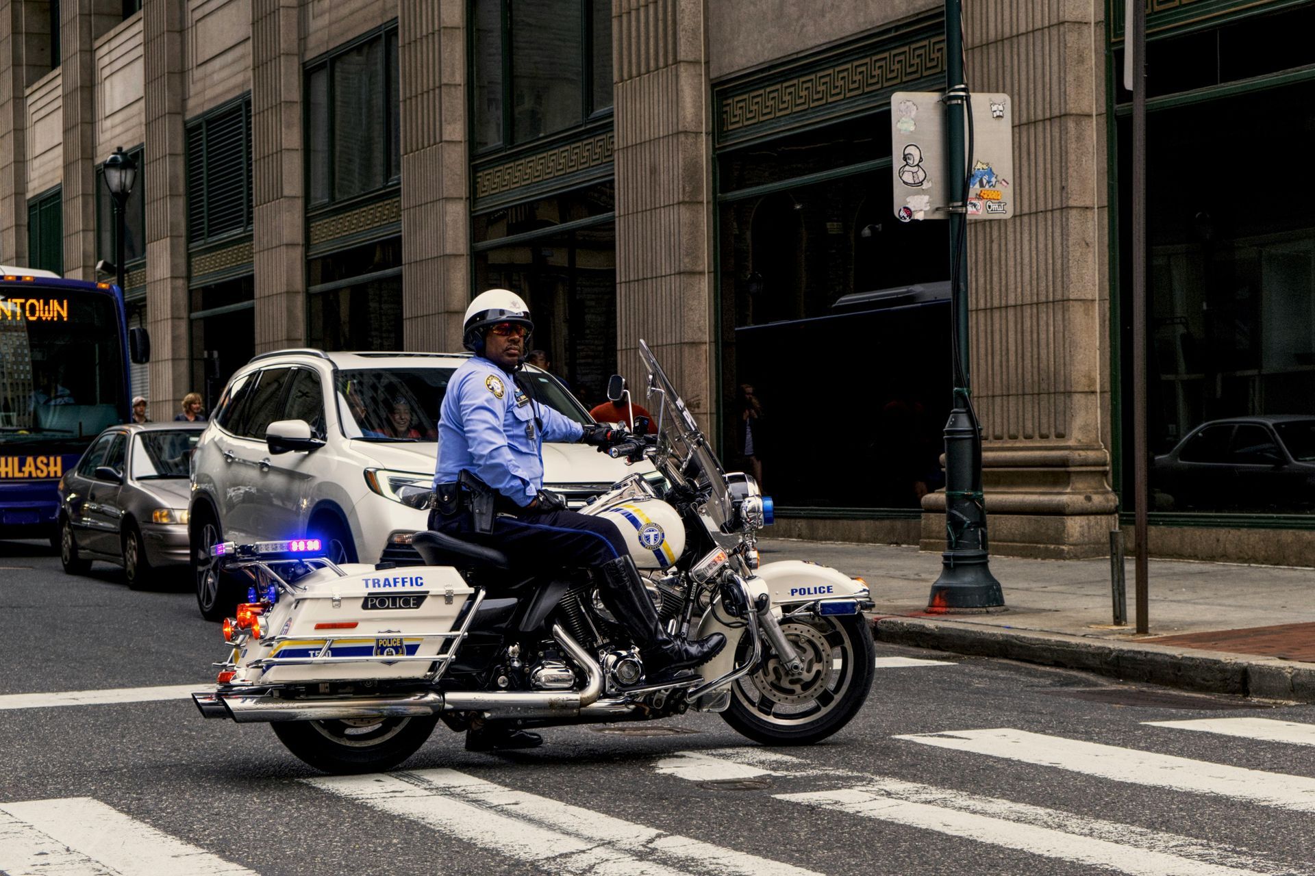 Police officer on a motorcycle in city traffic, blue and white bike, buildings in the background.