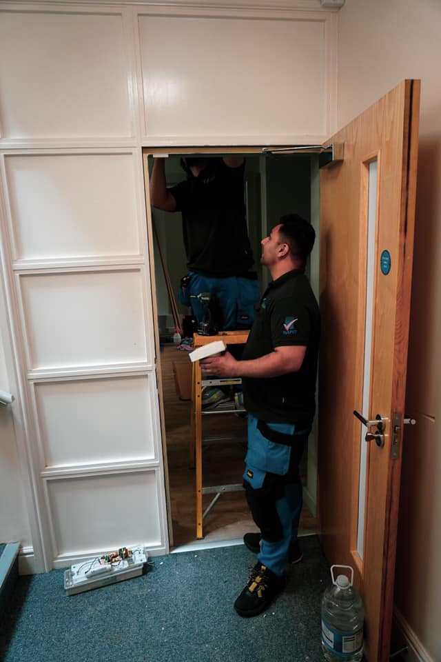 Two workers installing a door; one on a ladder, the other holding materials. White walls, wooden door, blue carpet.