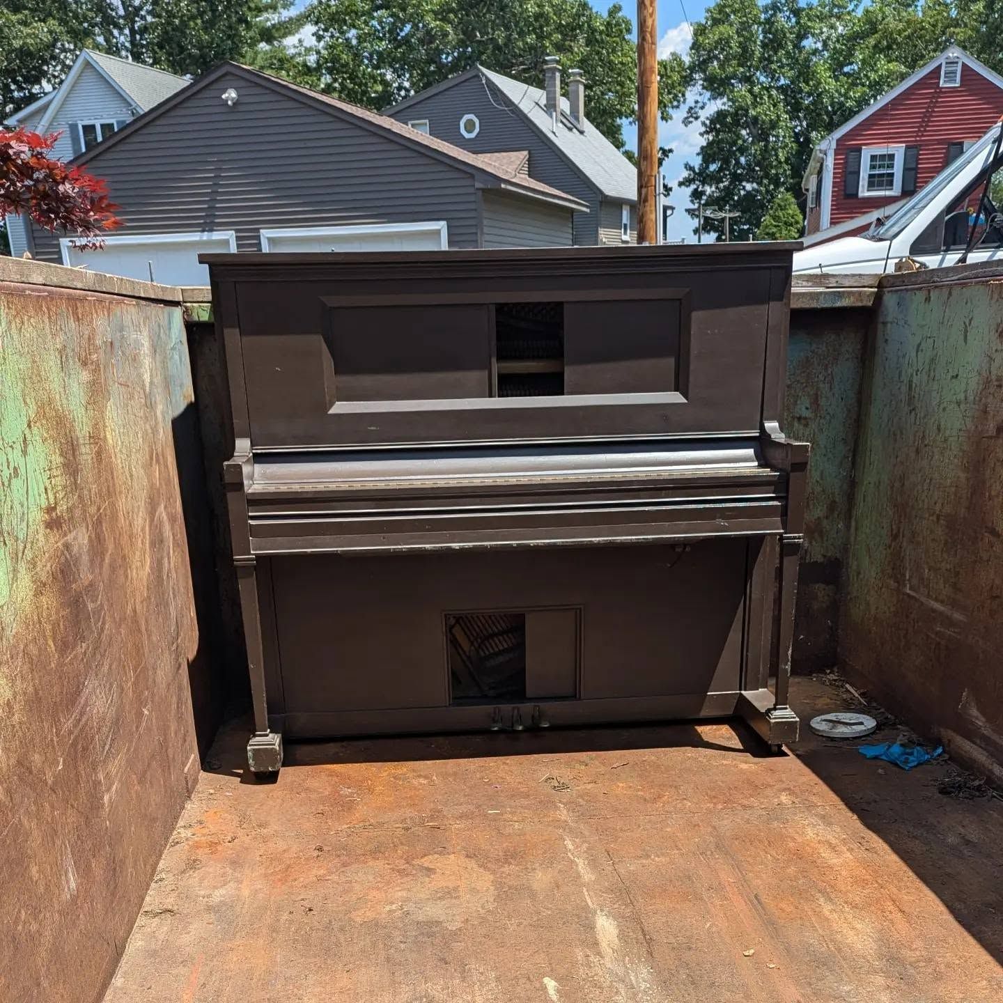 Old upright piano in a rusty metal dumpster, with houses visible in the background.