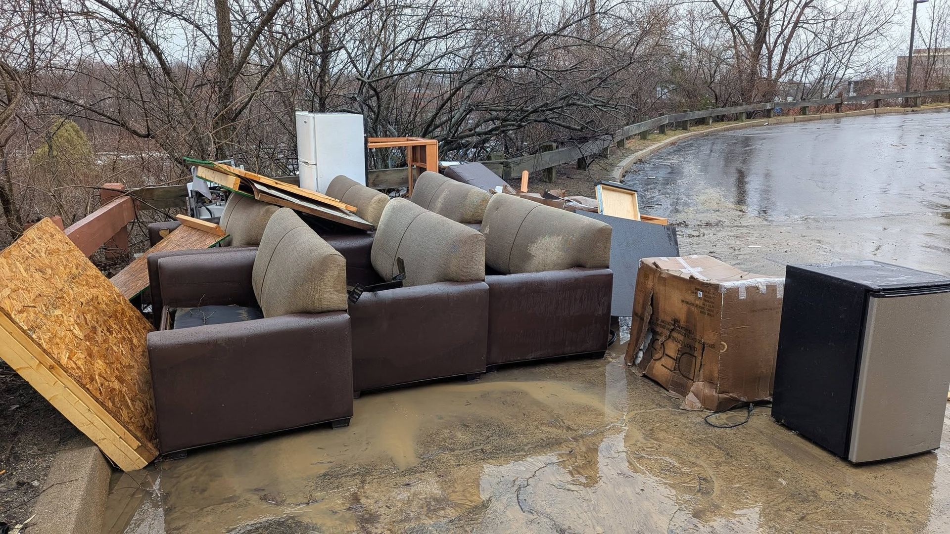 Flood-damaged outdoor furniture and debris piled beside a muddy riverbank, with trees in the background