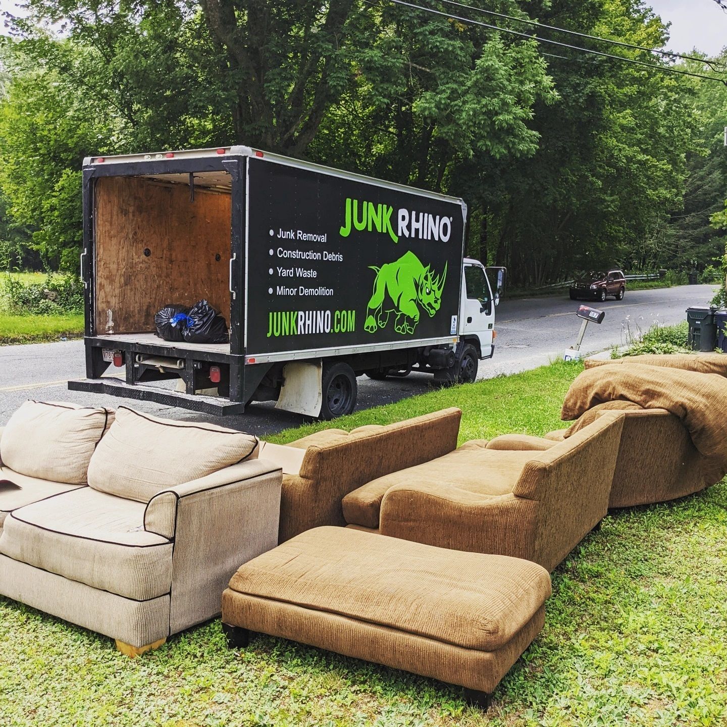 Junk removal truck with a logo next to old furniture on a lawn. A road and trees are in the background.