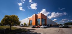Exterior view of a modern warehouse building with orange accents, parked cars, and a tree under a blue sky.