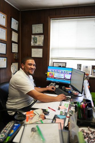 Man at an office desk smiling at a computer monitor, with papers and supplies in a wood-paneled office.