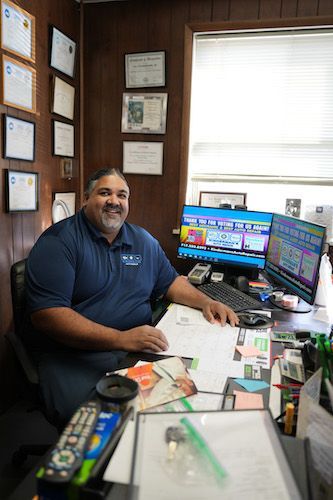 Man smiling at a cluttered office desk with dual monitors and papers near a bright window.