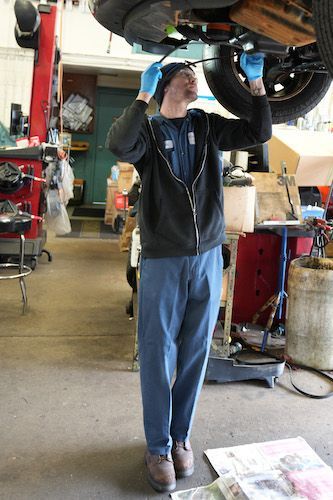 Mechanic working under a raised car in an auto repair shop, wearing blue gloves and dark clothes