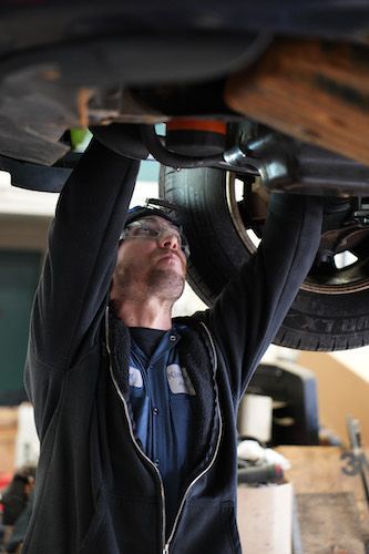 Mechanic working under a vehicle, inspecting or repairing the undercarriage in a garage.