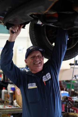 Mechanic smiling under a raised car in an auto shop, reaching up with both hands