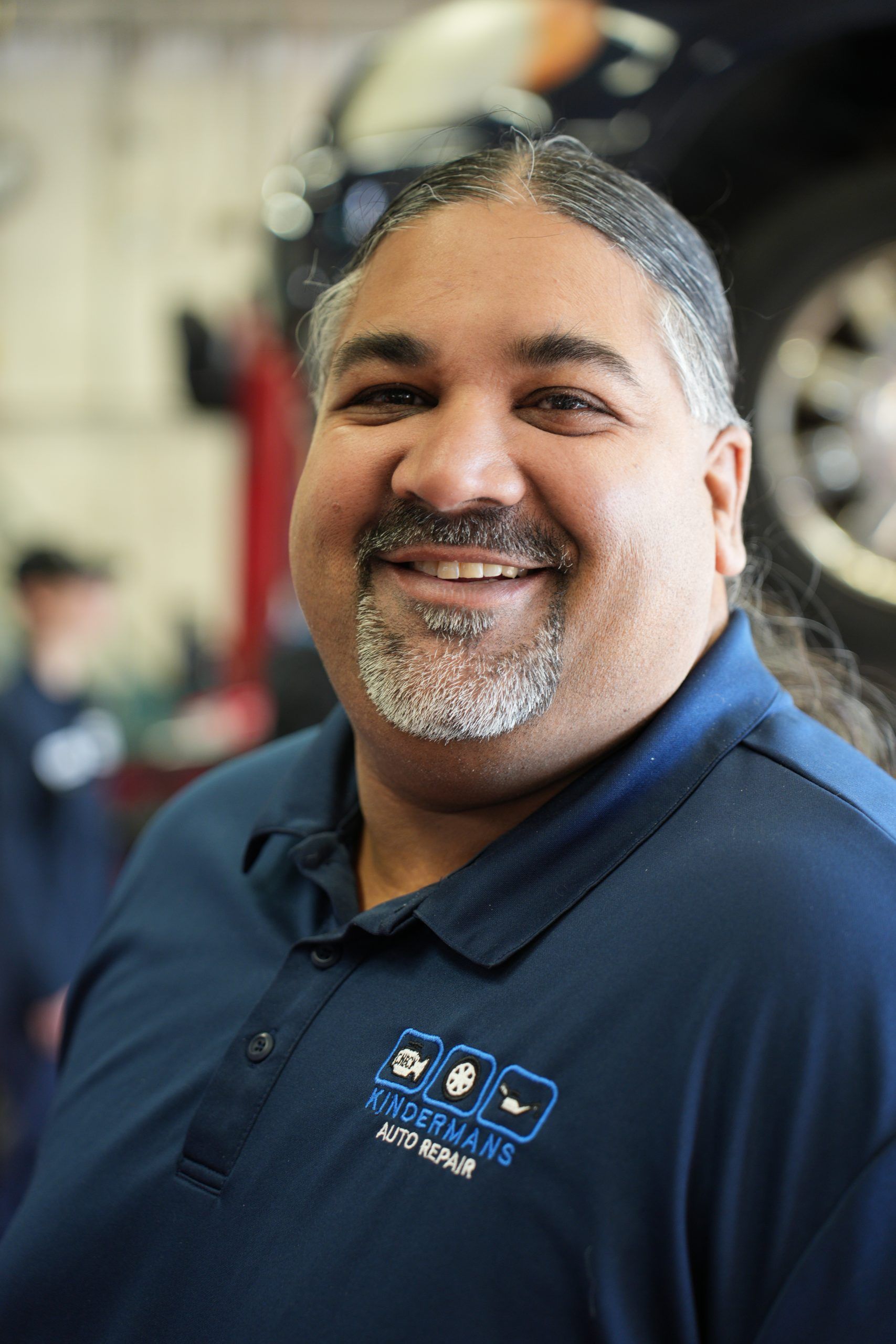 Smiling man in navy polo shirt standing in an auto shop, with a car tire blurred in the background