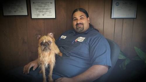 Man sitting with a small scruffy dog in a wood-paneled room, smiling at the camera.