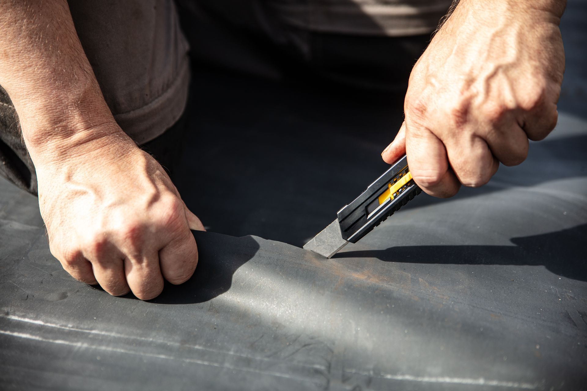 Hands use a utility knife to cut black roofing material on a flat surface.