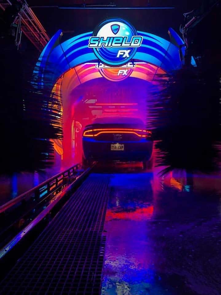 A car moving through a neon-lit, purple and red "Shield FX" car wash tunnel.