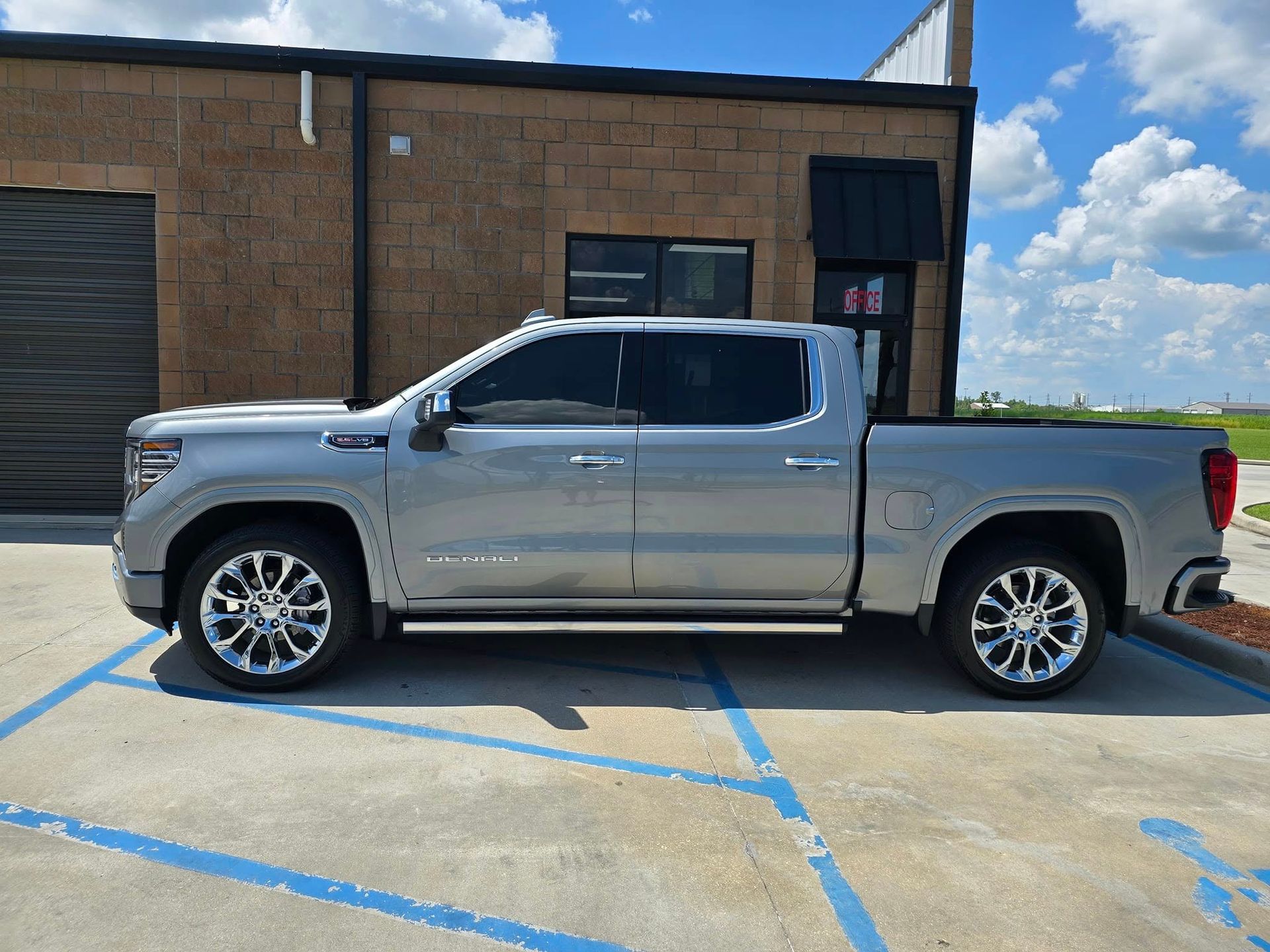 A silver crew cab pickup truck parked on a concrete lot in front of a tan brick building.