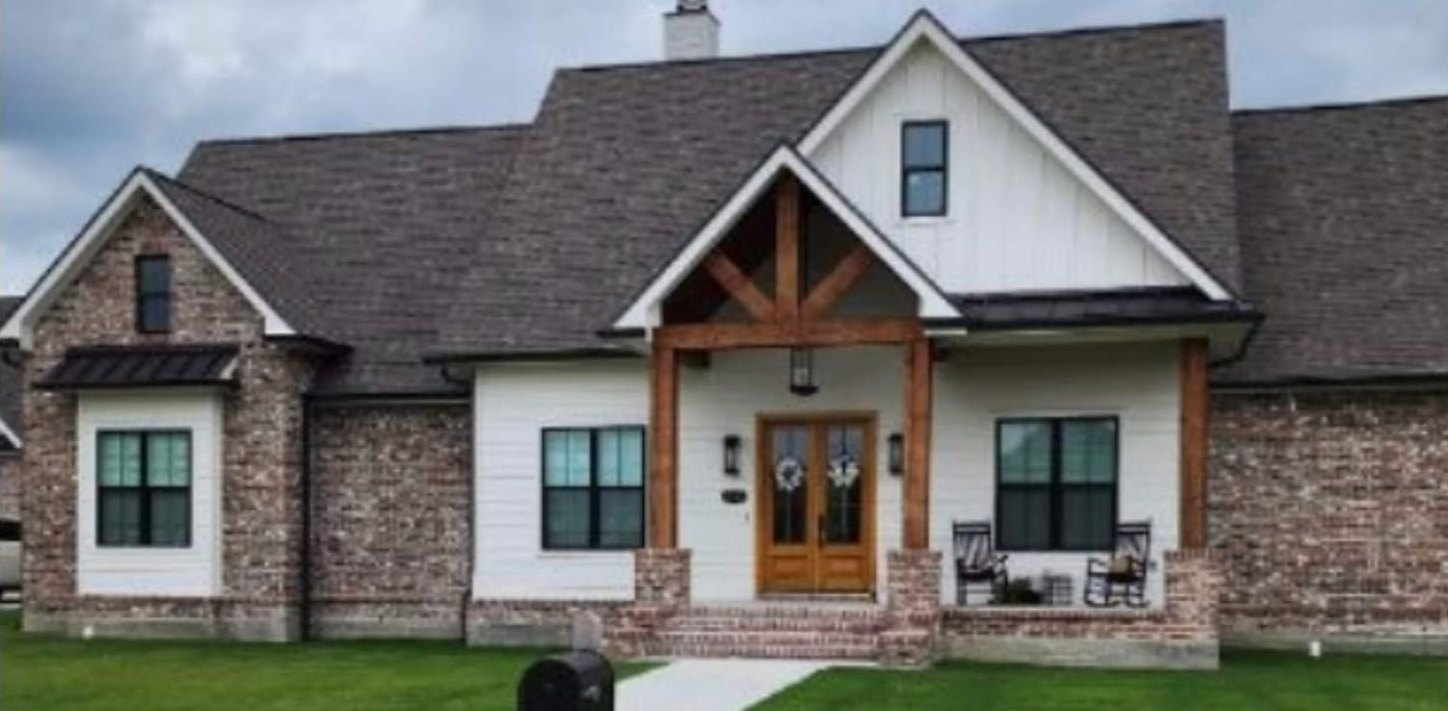 A single-story suburban house with white siding, brick accents, a gabled roof, and a wooden front porch entryway.