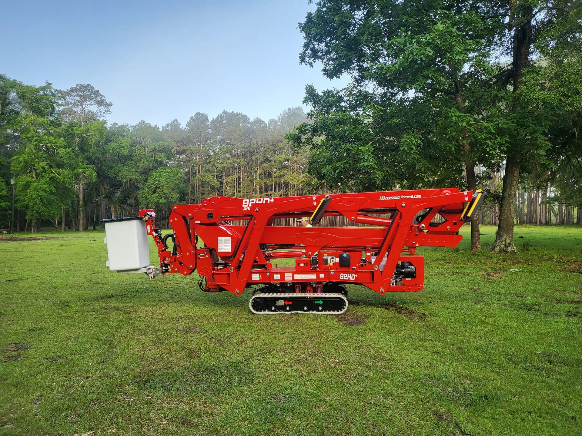 Red agricultural machine on grass in a park, with trees and a white box on the left.