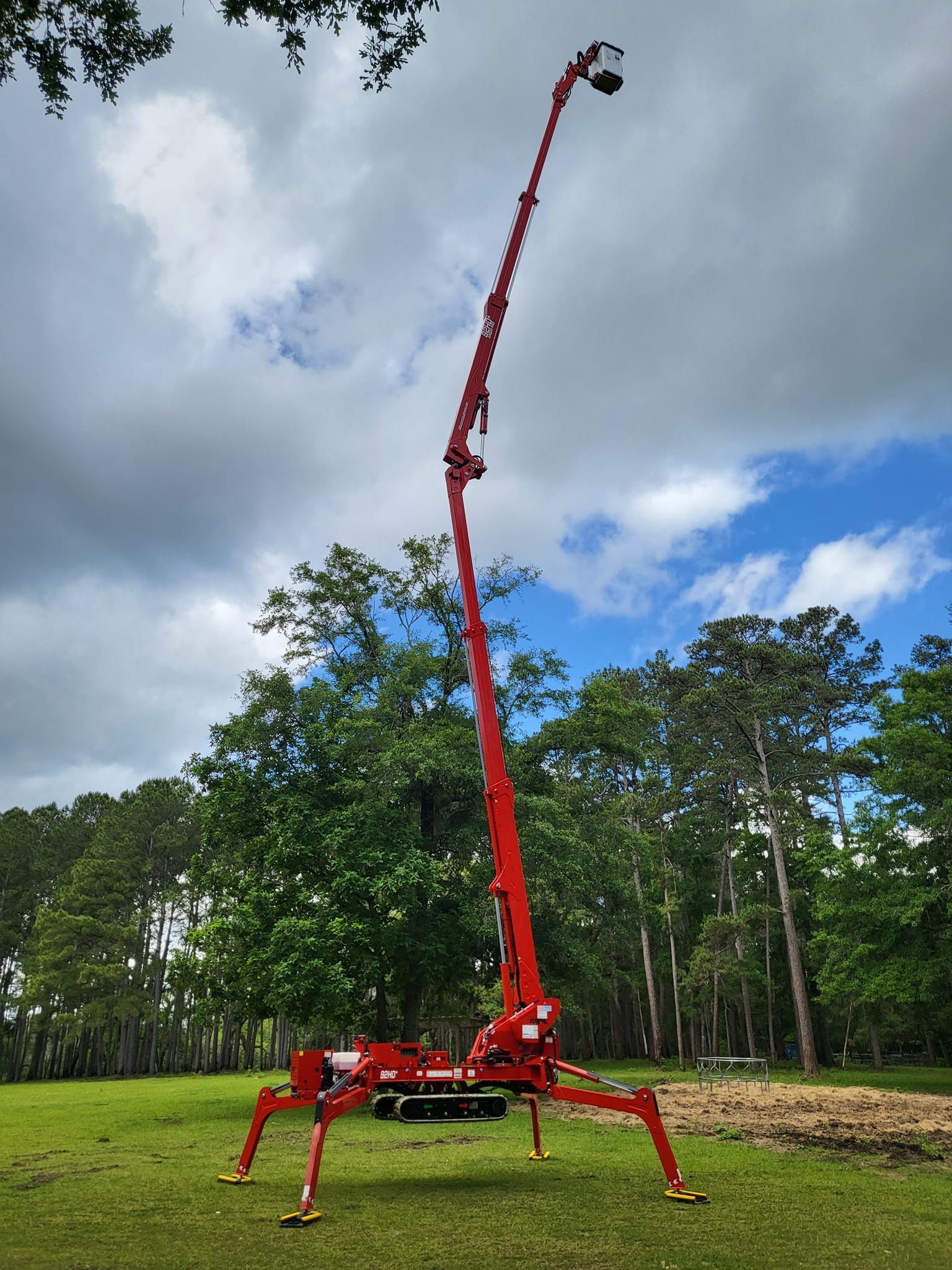 Red spider lift crane with a long boom raised high in a grassy field under cloudy sky.