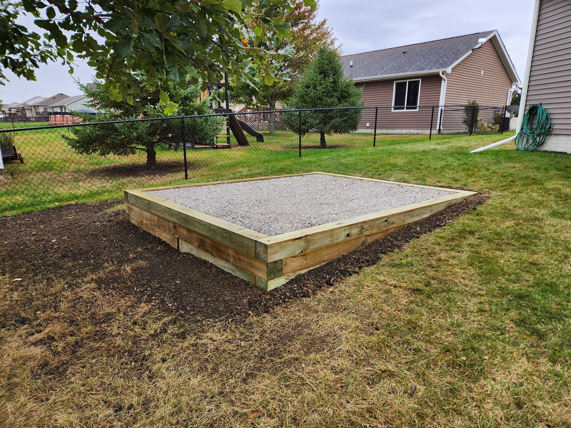 Raised wooden platform with gravel top in a grassy backyard, near a tree and a house
