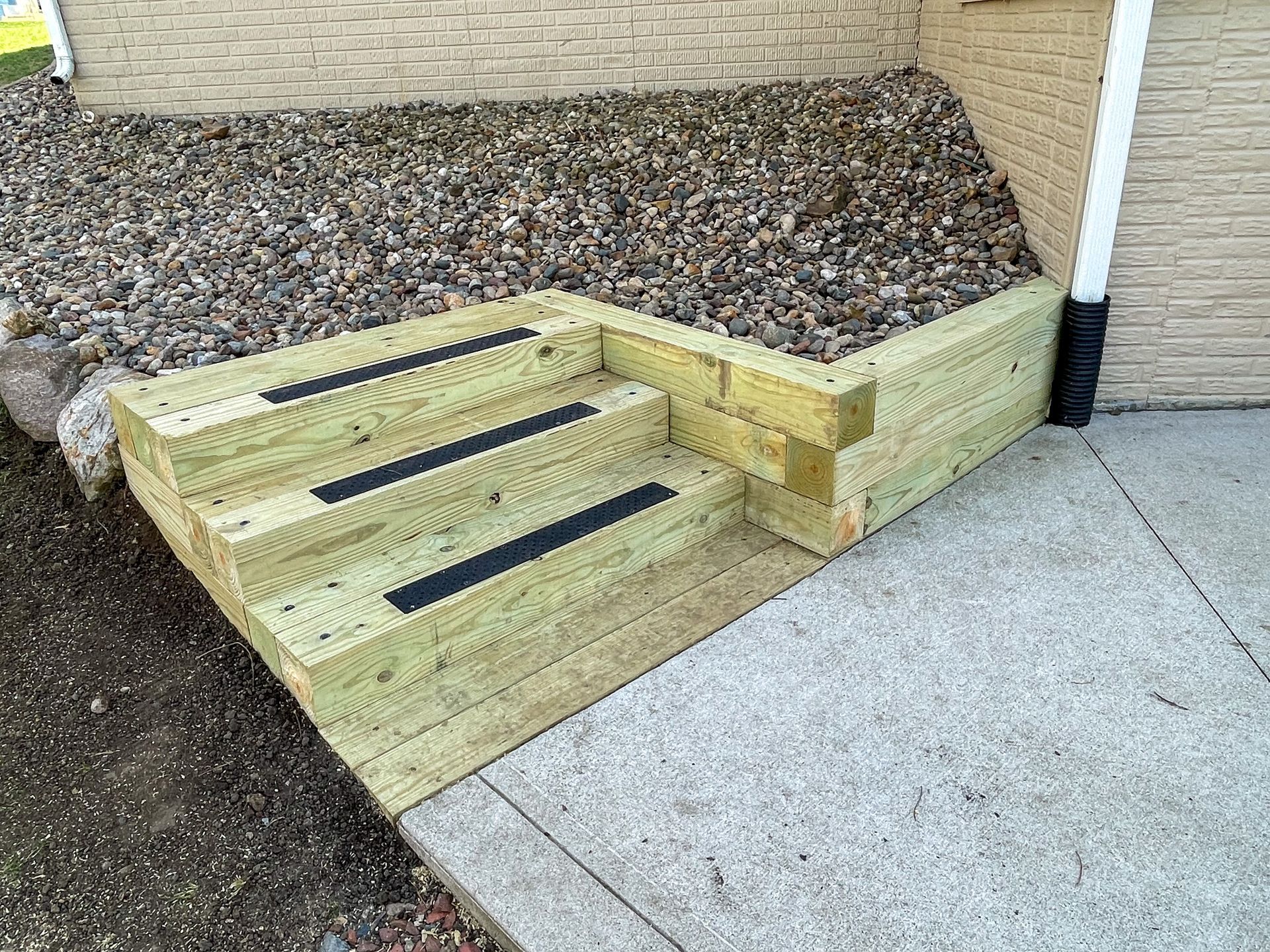 Wooden outdoor steps beside a concrete sidewalk and rock-covered slope against a brick wall
